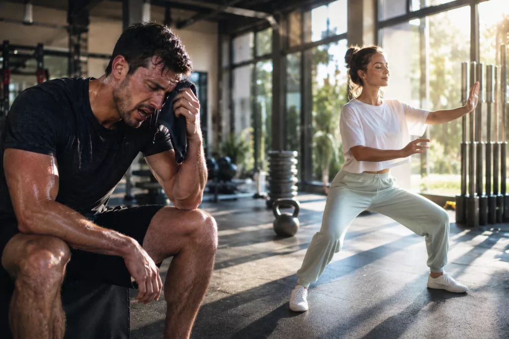 Dans une salle de sport pendant une période de forte chaleur, un sportif épuisé par l'effort contraste avec une pratiquante de Tai Chi calme et détendue, illustrant une approche plus douce du mouvement lors d'une canicule.